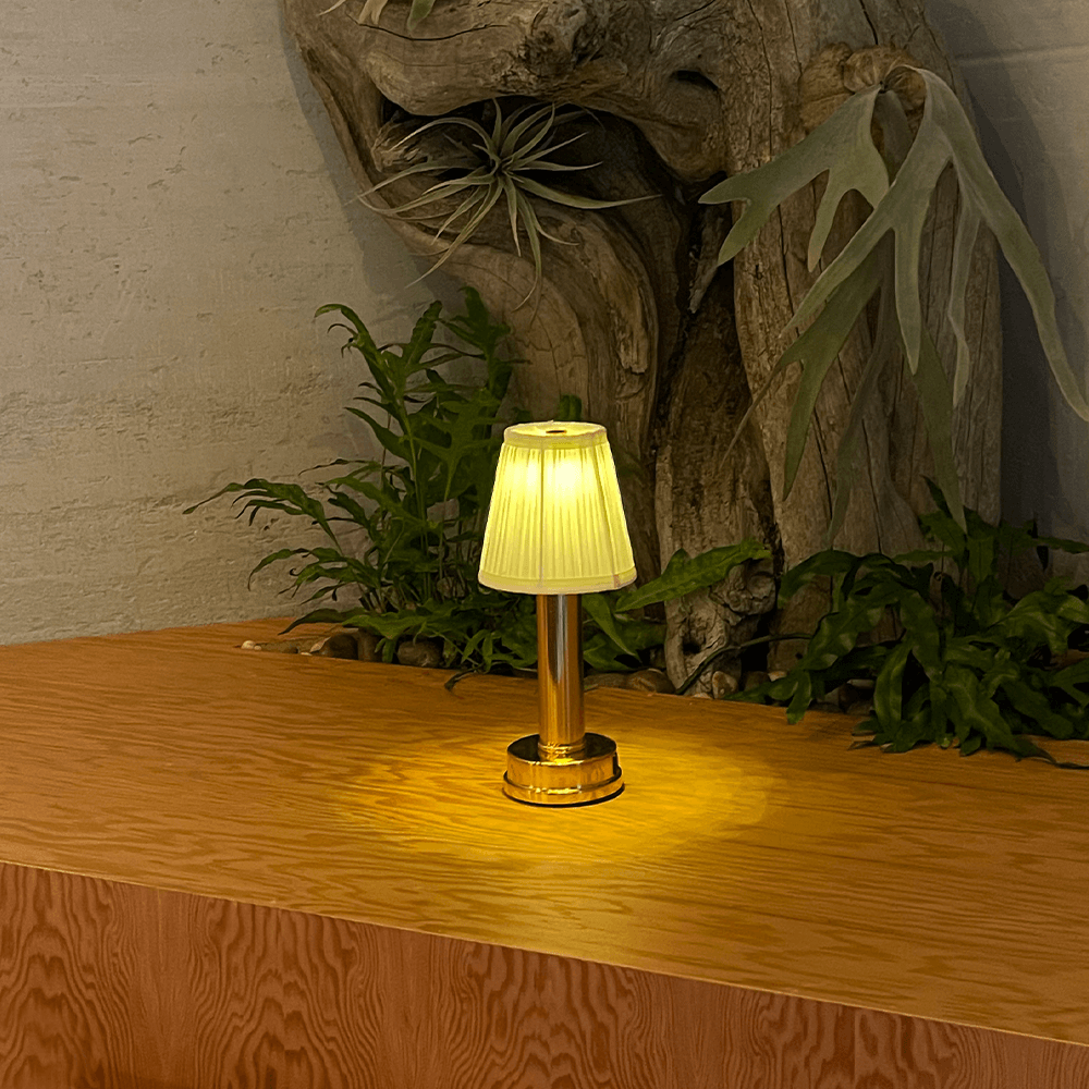 Gold cordless rechargeable touch table lamp lit on a wooden counter beside indoor plants