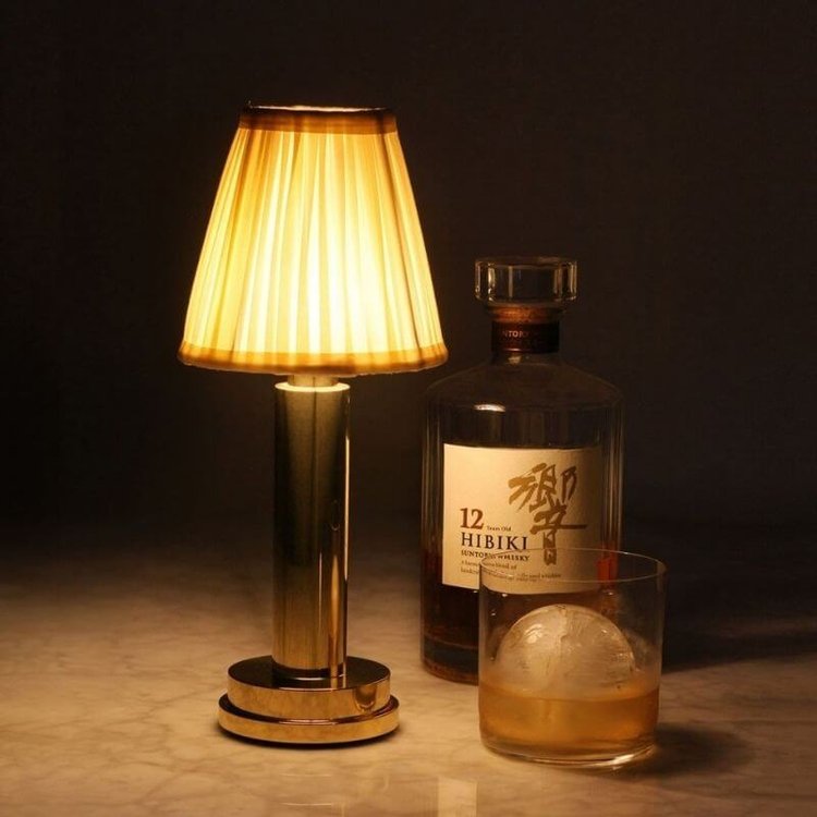 Gold cordless rechargeable touch table lamp lit on a bar top beside a whisky bottle and glass
